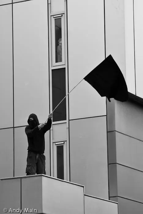 A Black Block member has scaled exchange tower and waves his flag tauntingly at police. Above an employee watches from the security of a high window.
